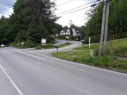 A gravel driveway leads to a house surrounded by trees, with a sign at the entrance and cars parked nearby.