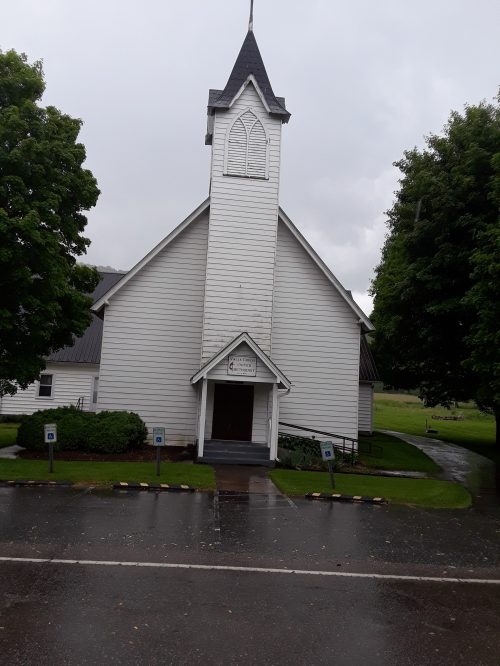 A white church with a tall steeple, surrounded by green trees, on a rainy day.