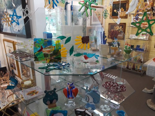 A display of colorful glass art pieces, including flowers, animals, and abstract shapes, arranged on a glass shelf.