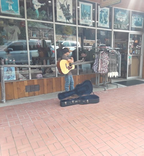 A young musician plays guitar outside a shop, with a guitar case open for tips and clothing displayed in the window.