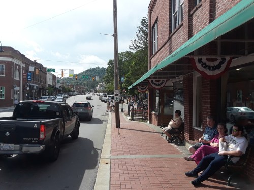 A street view of a small town with shops, parked cars, and people sitting on benches under awnings.