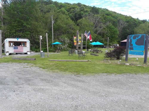 A cozy outdoor area with picnic tables, a food truck, and flags, surrounded by greenery and a clear blue sky.