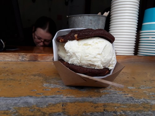 A chocolate cookie sandwich filled with vanilla ice cream, resting on a wooden counter. A person is blurred in the background.