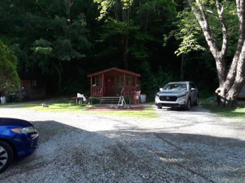 A small red cabin surrounded by trees, with a gravel driveway and parked cars nearby.