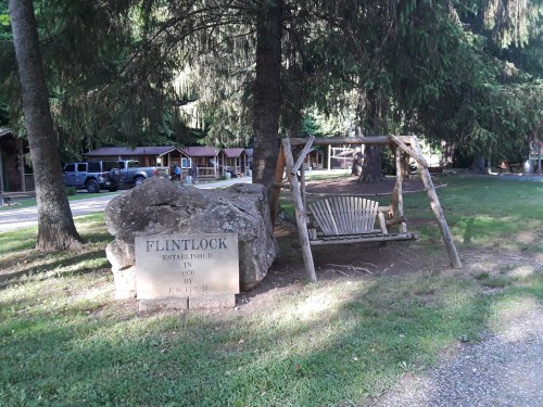 A wooden swing next to a large rock sign reading "Flintlock Established in 1870" surrounded by trees.