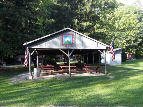 A covered picnic area with benches, American flags, and a mountain-themed decoration, surrounded by greenery.