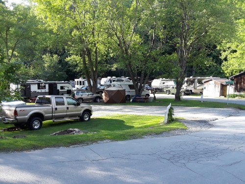 A parking area with several RVs and a pickup truck surrounded by trees and green grass.