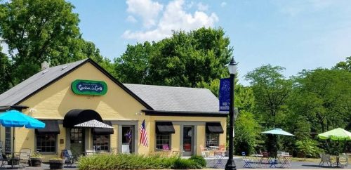 A charming café with outdoor seating, surrounded by greenery and colorful umbrellas under a blue sky.