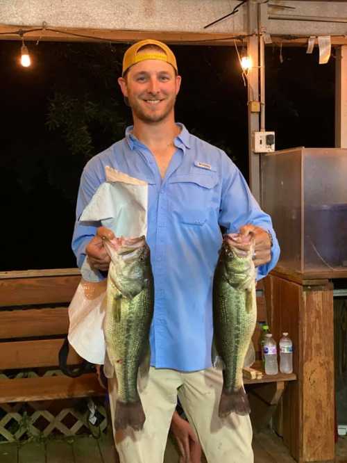 A smiling man in a blue shirt holds two large bass fish, standing in a rustic setting with warm lighting.