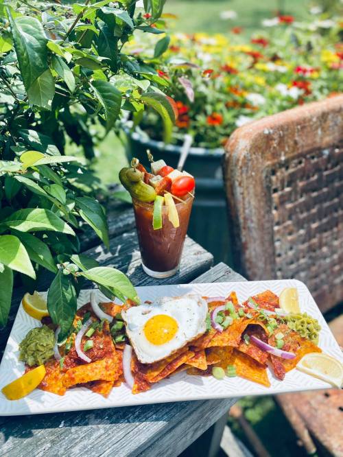 A plate of nachos topped with an egg, served with a colorful Bloody Mary, surrounded by vibrant flowers.