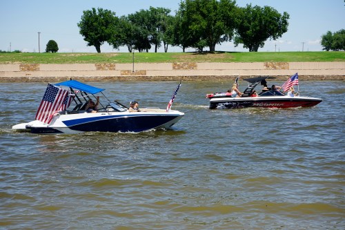 Two boats with American flags cruising on a lake, surrounded by trees and a clear blue sky.