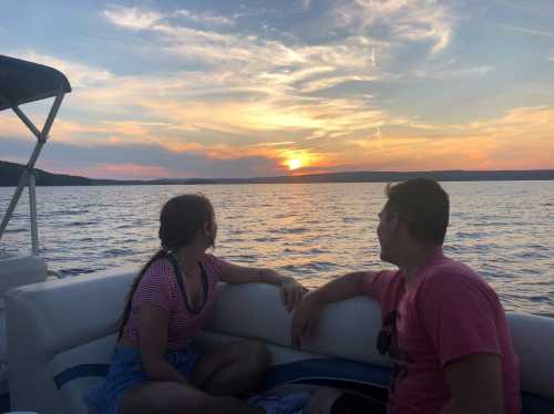 A couple sits on a boat, watching a vibrant sunset over the water, with colorful clouds in the sky.