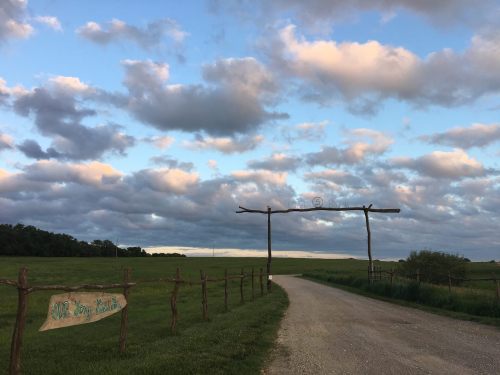 A gravel road leads through a grassy field under a sky filled with fluffy clouds, marked by a wooden signpost.