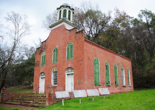 Historic red brick building with green shutters, surrounded by trees, featuring a small cupola on the roof.