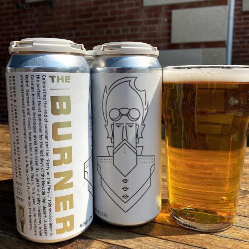 Two cans of "The Burner" beer beside a glass of golden beer on a wooden table, with a brick wall in the background.