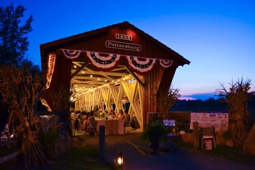 A rustic covered bridge lit up for an evening event, decorated with bunting, surrounded by a serene twilight landscape.