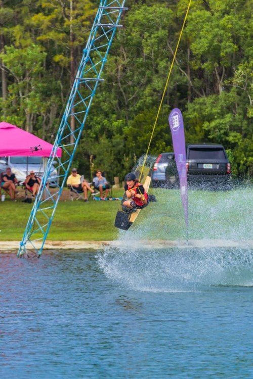 A person jumps off a wakeboard while being towed over a lake, with spectators and a pink tent in the background.