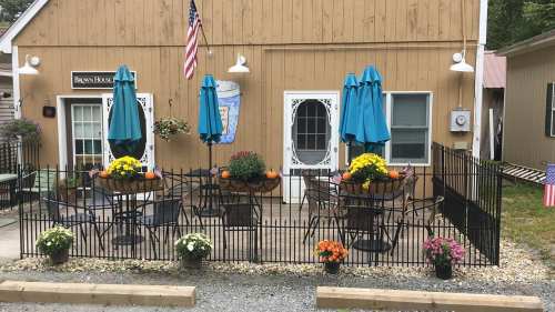 A cozy outdoor seating area with tables, blue umbrellas, and colorful flower pots, surrounded by a black fence.