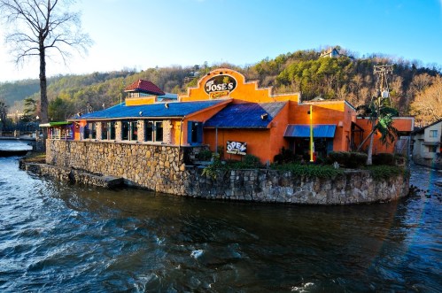 Colorful restaurant on a waterfront, surrounded by trees and hills, with a blue sky above.