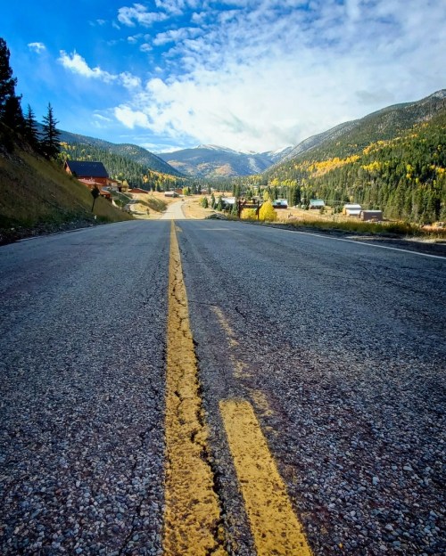 A wide, empty road stretches into the distance, flanked by mountains and trees under a blue sky with scattered clouds.