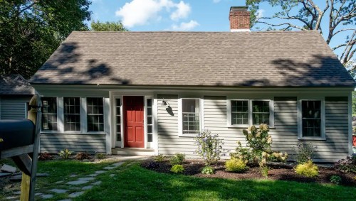 A charming gray house with a red door, surrounded by greenery and a clear blue sky.