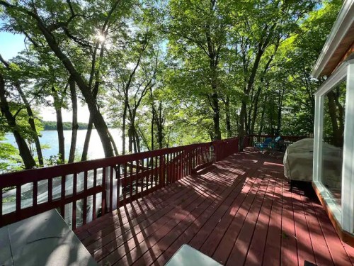 A wooden deck surrounded by trees, overlooking a serene lake with sunlight filtering through the leaves.