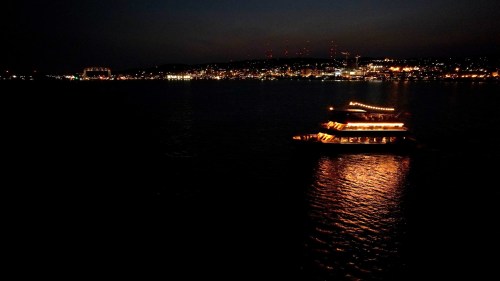 Boats with lights glide across a dark waterway, reflecting city lights in the background at night.