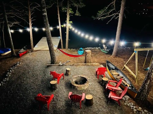 A cozy campsite at night with a fire pit, red chairs, a hammock, and string lights by a lake.