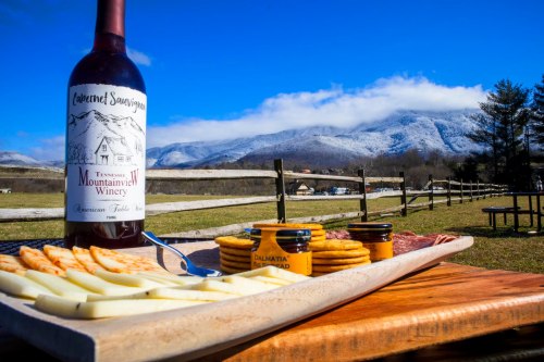 A scenic outdoor setting featuring a bottle of wine, cheese, crackers, and mustard, with snow-capped mountains in the background.