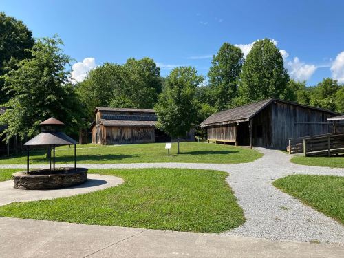 A sunny outdoor scene featuring rustic wooden buildings, a grassy area, and a stone well surrounded by trees.