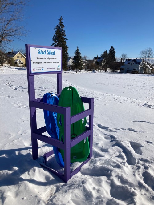 A colorful sled rack in a snowy field, featuring blue and green sleds with a sign encouraging sharing.