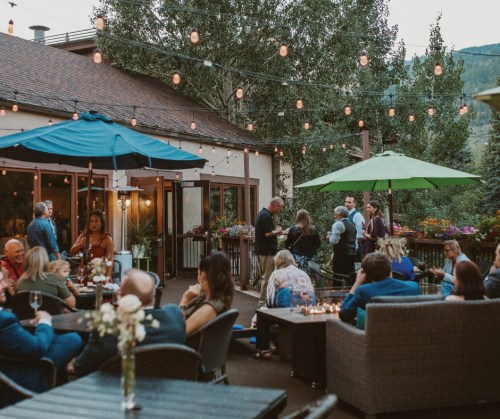 A lively outdoor gathering with people socializing, string lights, and umbrellas at a scenic venue.