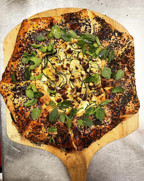 A freshly baked pizza on a wooden peel, topped with herbs and black sesame seeds, with a golden, crispy crust.