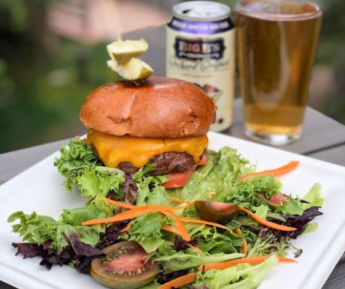 A juicy cheeseburger on a bun with a pickle, served with a fresh salad and a can of beer in the background.