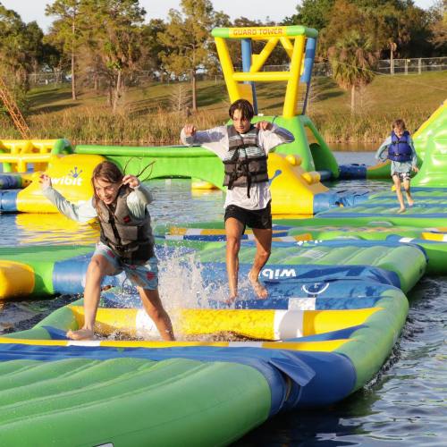 Two children play on a colorful water obstacle course, splashing as they run across inflatable platforms.