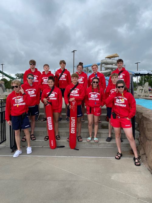 A group of lifeguards in red uniforms poses together by a pool, with water slides in the background.