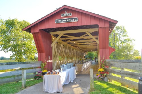 A red covered bridge with string lights, leading to a decorated outdoor event space with tables and flowers.