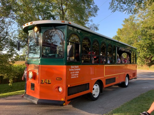A colorful trolley bus with orange and green colors parked on a tree-lined road, filled with passengers.