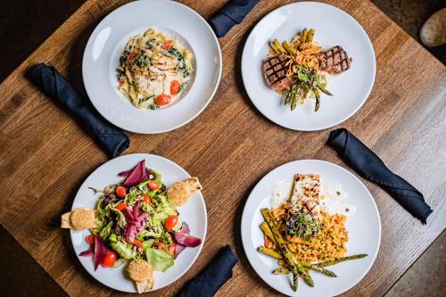 Four plates of gourmet dishes on a wooden table, featuring pasta, grilled meat, salad, and vegetables.