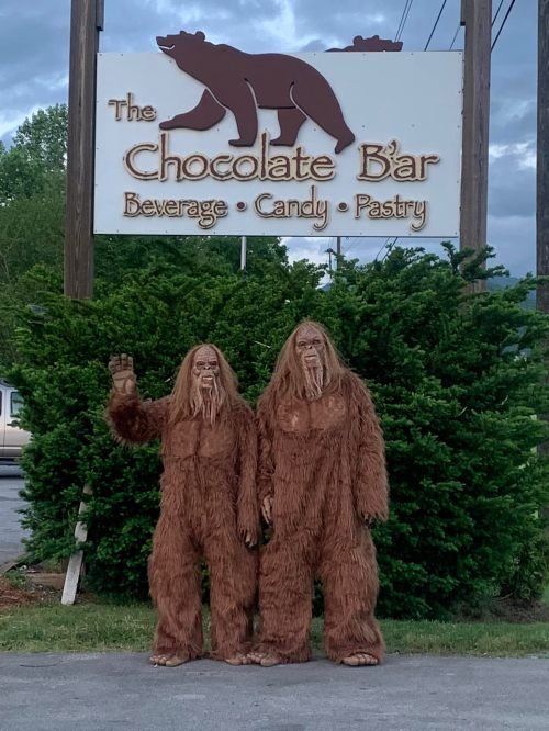 Two people in Bigfoot costumes stand in front of a sign for "The Chocolate B'ar" with trees in the background.