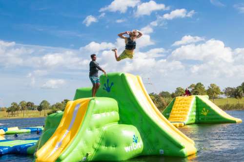 A person jumps off an inflatable water slide while another stands on it, with a sunny sky and water in the background.