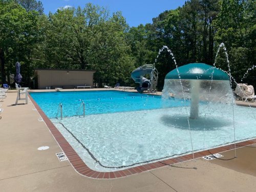 A sunny outdoor pool with a water fountain, slide, and lounge chairs surrounded by trees.