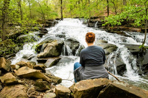 A person sits on rocks by a waterfall, surrounded by lush greenery and flowing water.