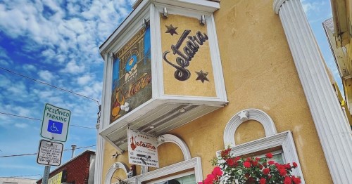 A colorful building with a sign reading "Gelateria" and flowers below, set against a blue sky with clouds.