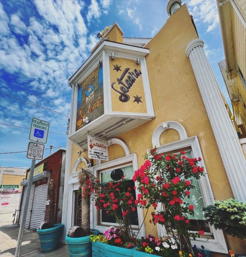 A colorful building with flower pots, featuring a sign that reads "Gelato" under a bright blue sky with clouds.
