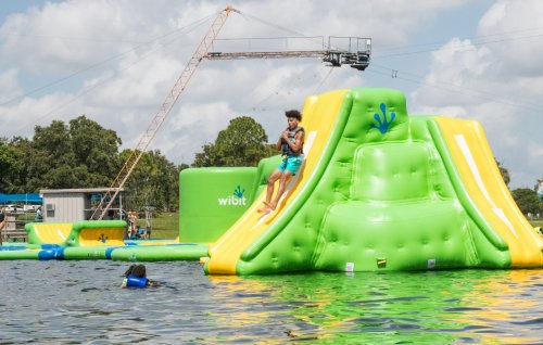A bright green inflatable water slide with a person sliding down, surrounded by a lake and another person swimming nearby.