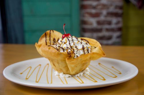 A dessert in a crispy bowl, topped with whipped cream, chocolate drizzle, and a cherry, served on a white plate.