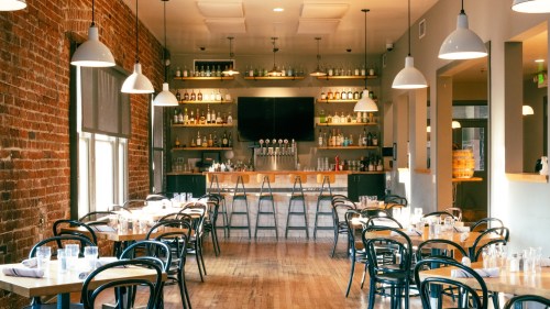 A cozy restaurant interior with wooden floors, brick walls, and tables set for dining, featuring a bar in the background.
