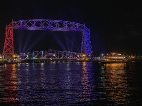 A brightly lit bridge at night, reflecting colorful lights on the water below.