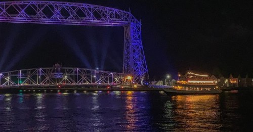 A brightly lit bridge at night, with colorful lights reflecting on the water and a boat passing by.
